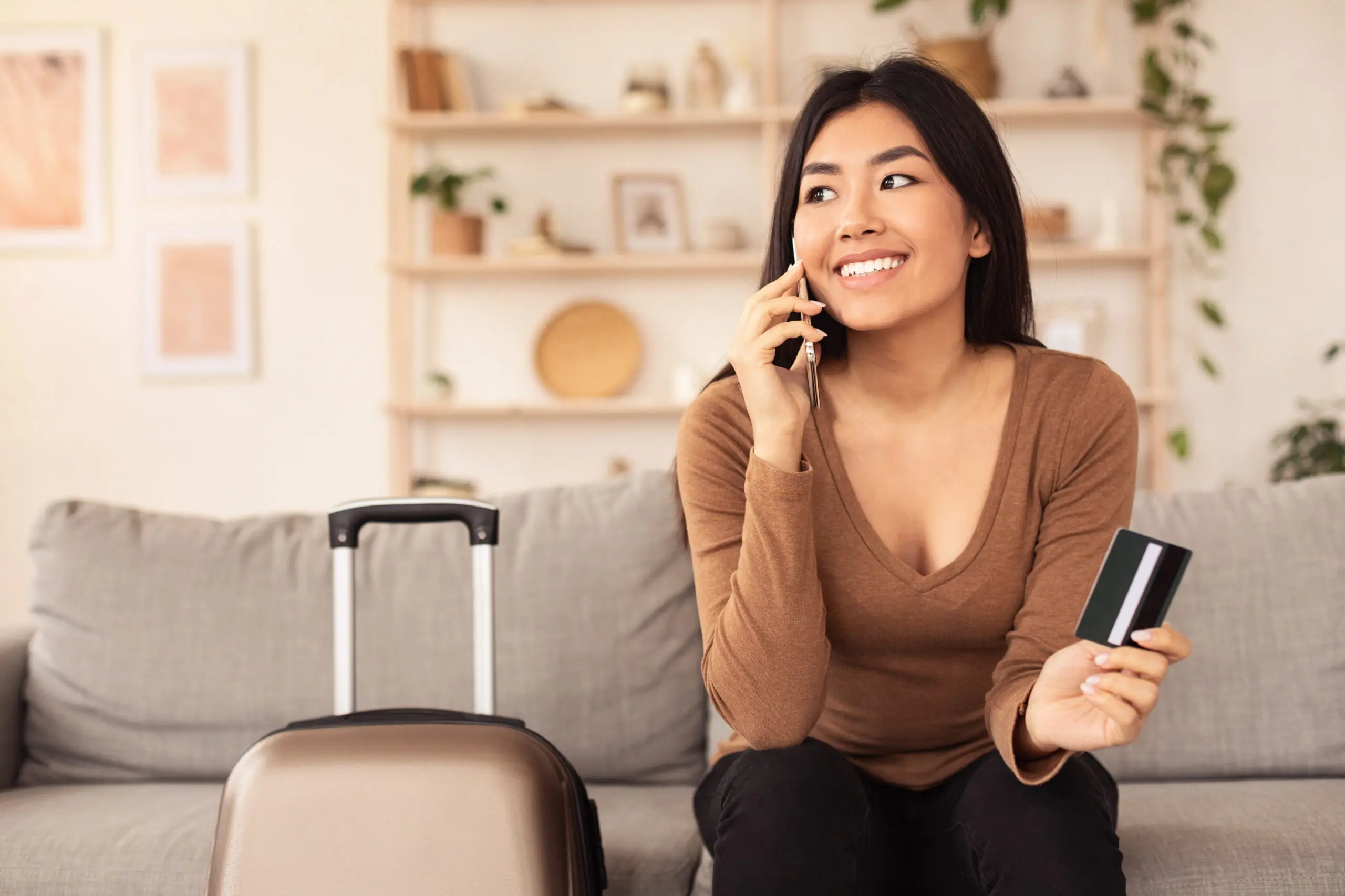 Transfer To Airport. Smiling Girl Ordering Taxi By Phone Holding Credit Card Sitting With Travel Suitcase At Home