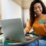 Fast Online Shopping. Smiling young black lady holding and looking at debit credit card using pc, customer making financial transaction sitting on chair, selective focus on hand, blurred background