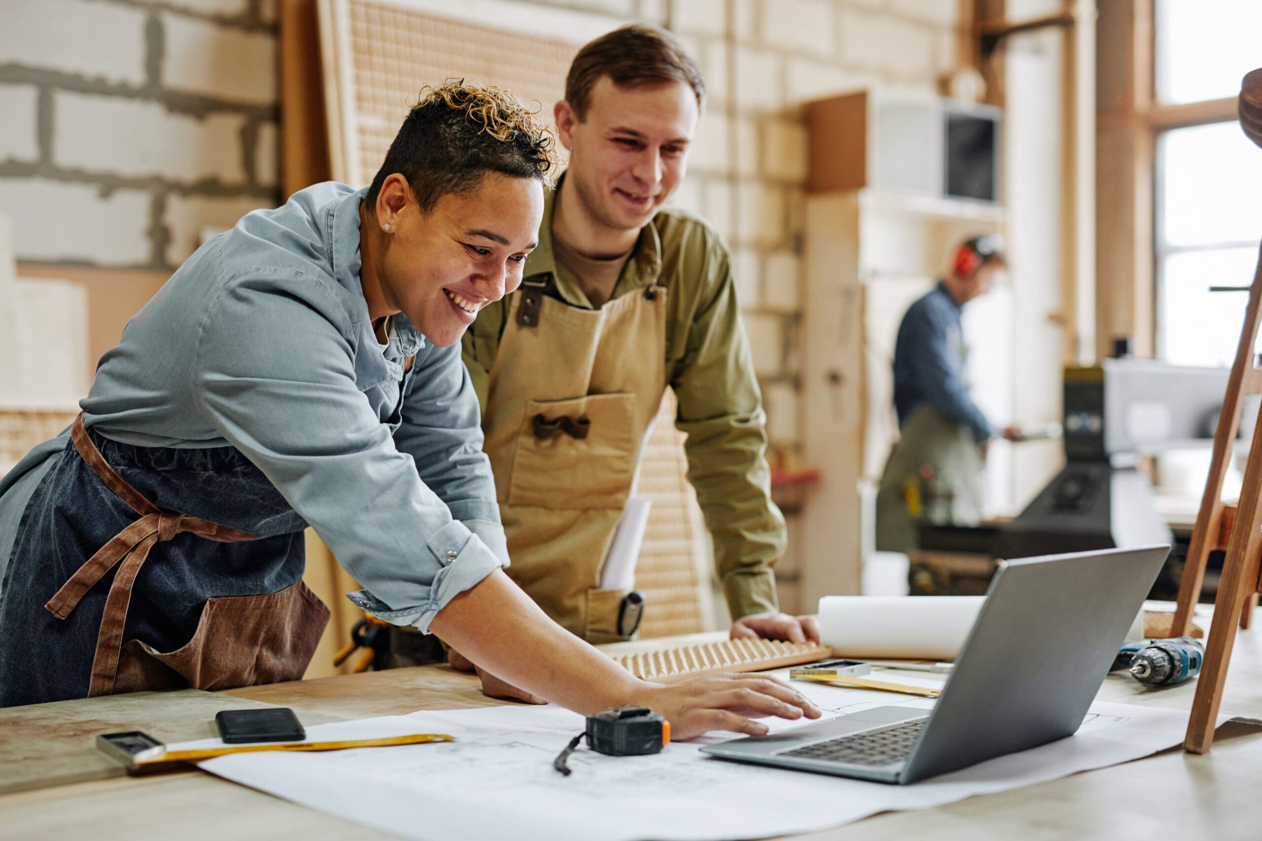 Warm toned portrait of animated carpenters using laptop and discussing furniture designs in workshop