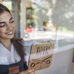 Portrait shot of a local business owner hanging an open sign on the retail glass window. Smiling female barber hanging open sign on door. Female owner is holding OPEN sign seen through glass.