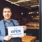 Small business owner smiling while holding open sign of restaurant reopening after lockdown quarantine due to coronavirus pandemic - Entrepeneur opening cafeteria activity to support local businesses.