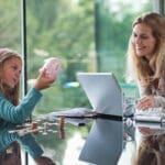 Mother Watching Young Daughter With Piggy Bank Counting Coins At A Glass Table