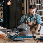 Young bearded carpenter focused on planning woodworking project using laptop in workshop