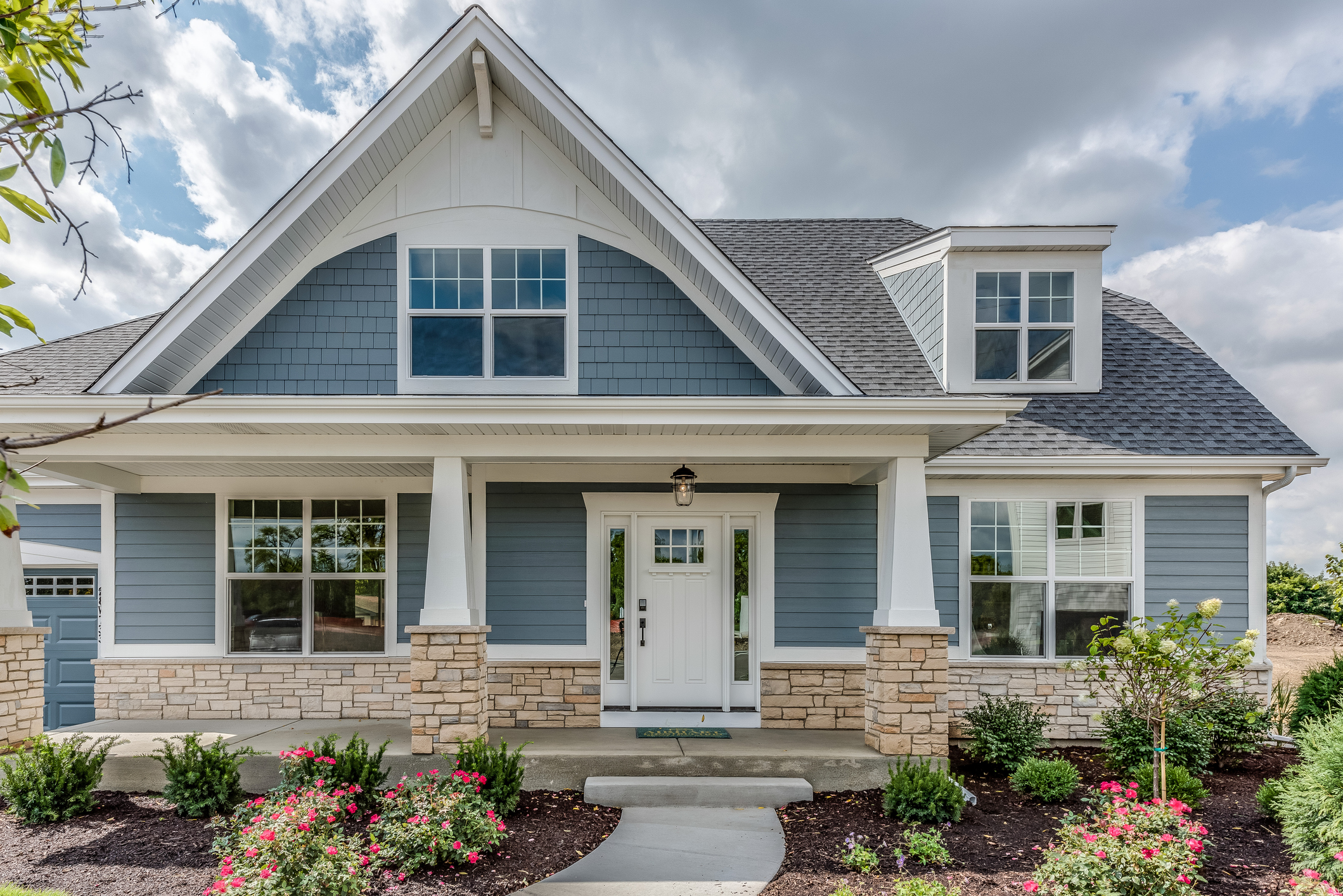 A large modern light blue house with a grey roof with lots of shrubs in beautifully landscaped yard