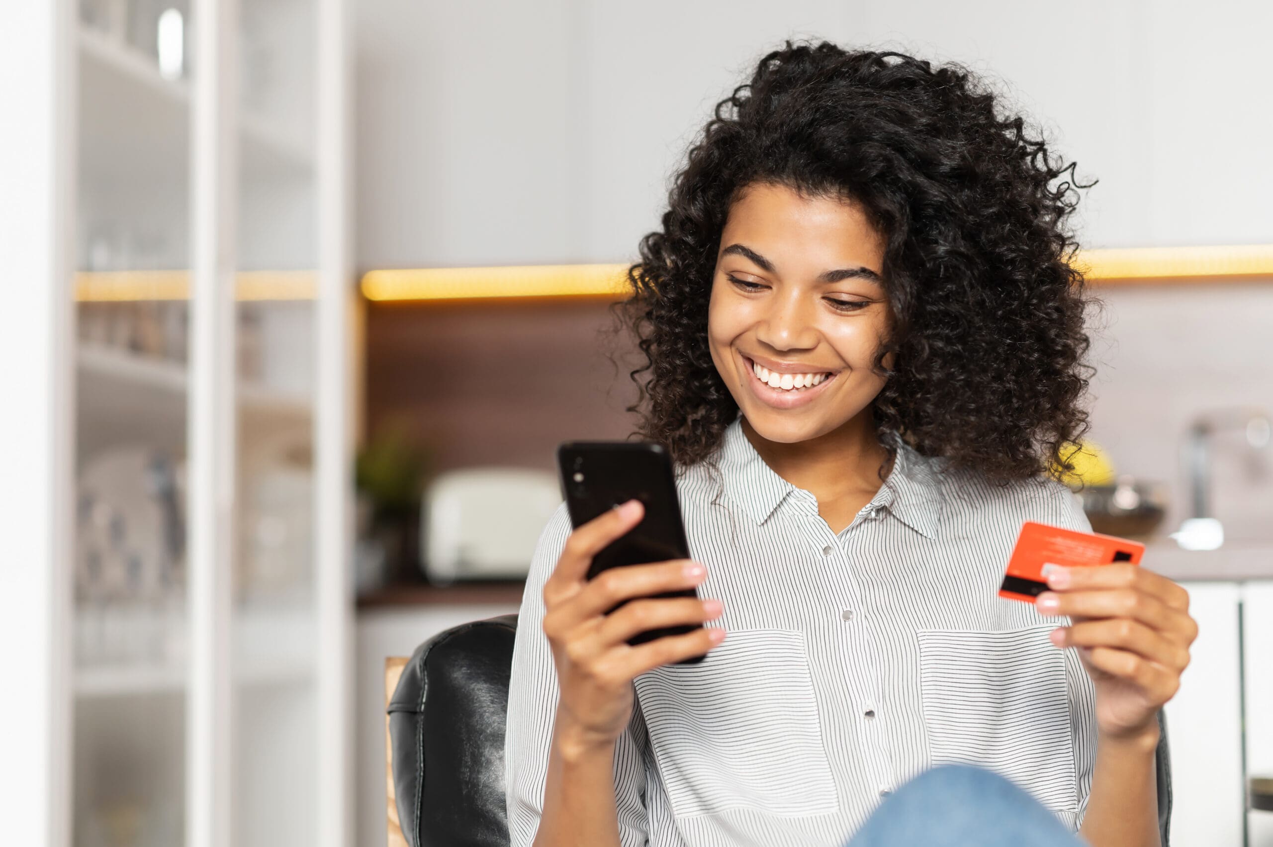 Smiling African American teenage girl with curly hair holding mobile phone, entering credit card number to make an online transaction, mixed-race woman ordering food, doing online shopping from home