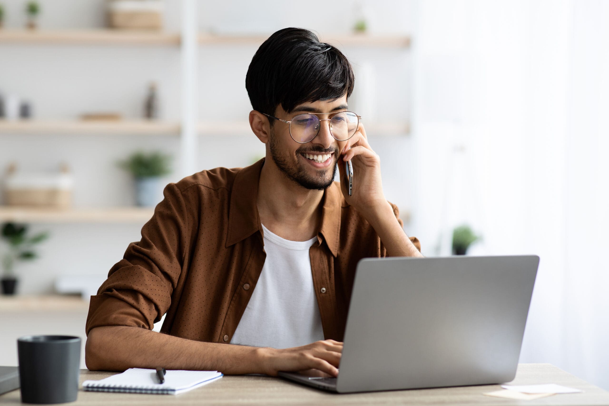 Cheerful indian guy businessman sitting at workdesk with laptop having phone conversation with business partner or client, typing on computer keyboard, office interior, copy space