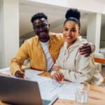 Portrait of a diverse couple sitting at the table at home and using a laptop and a house plan while making a financial strategy for building a new home. Looking in the camera. Moving in together.