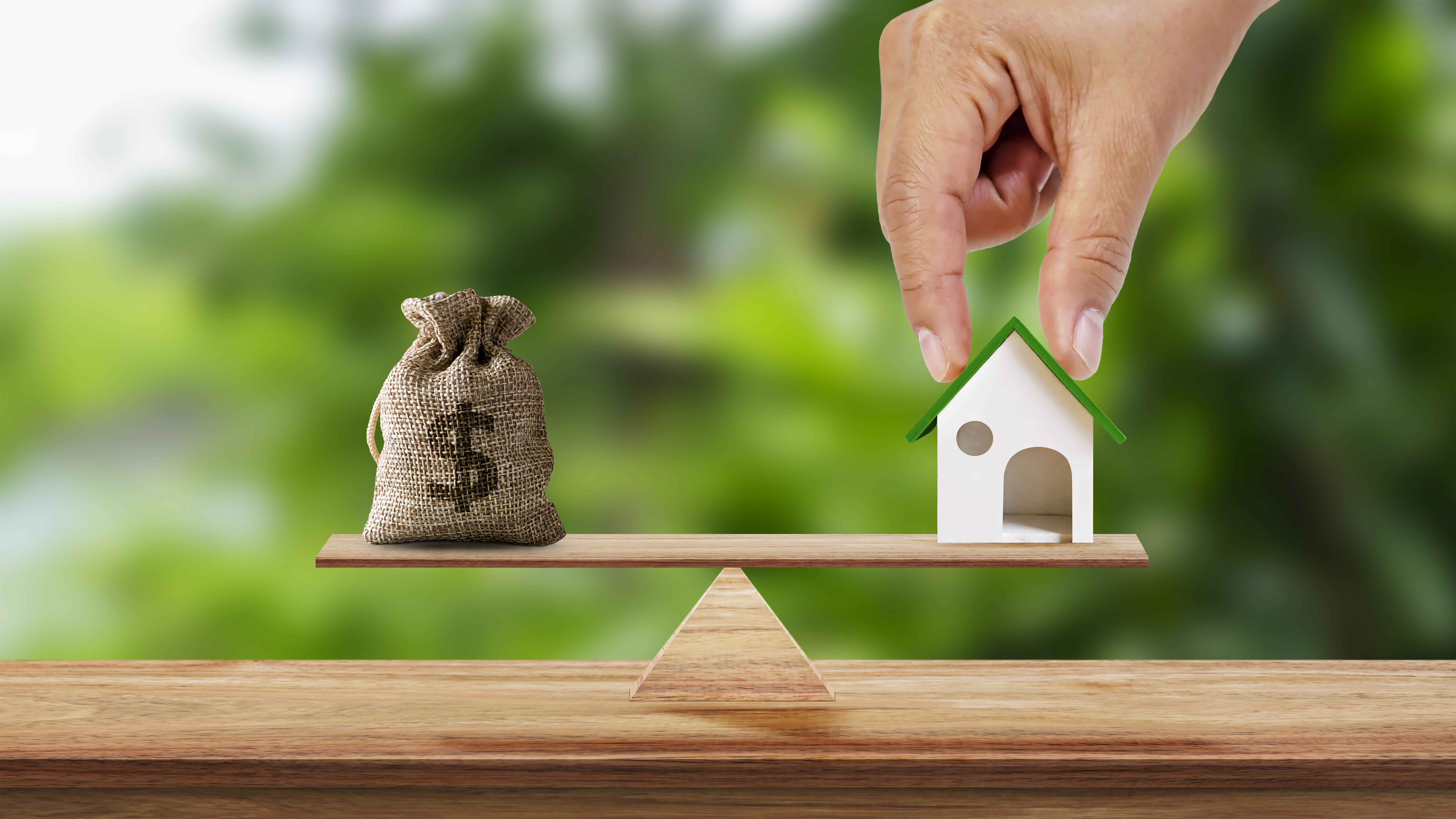 Hand holding model of house and savings bag placed on scales and blurred green background. Real estate ideas and future investment business plans.