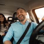 Nice Ride. Portrait of happy male driver riding car looking at cheerful beautiful lady sitting inside auto on back passenger seat, female using cell phone and talking with guy, windshield view