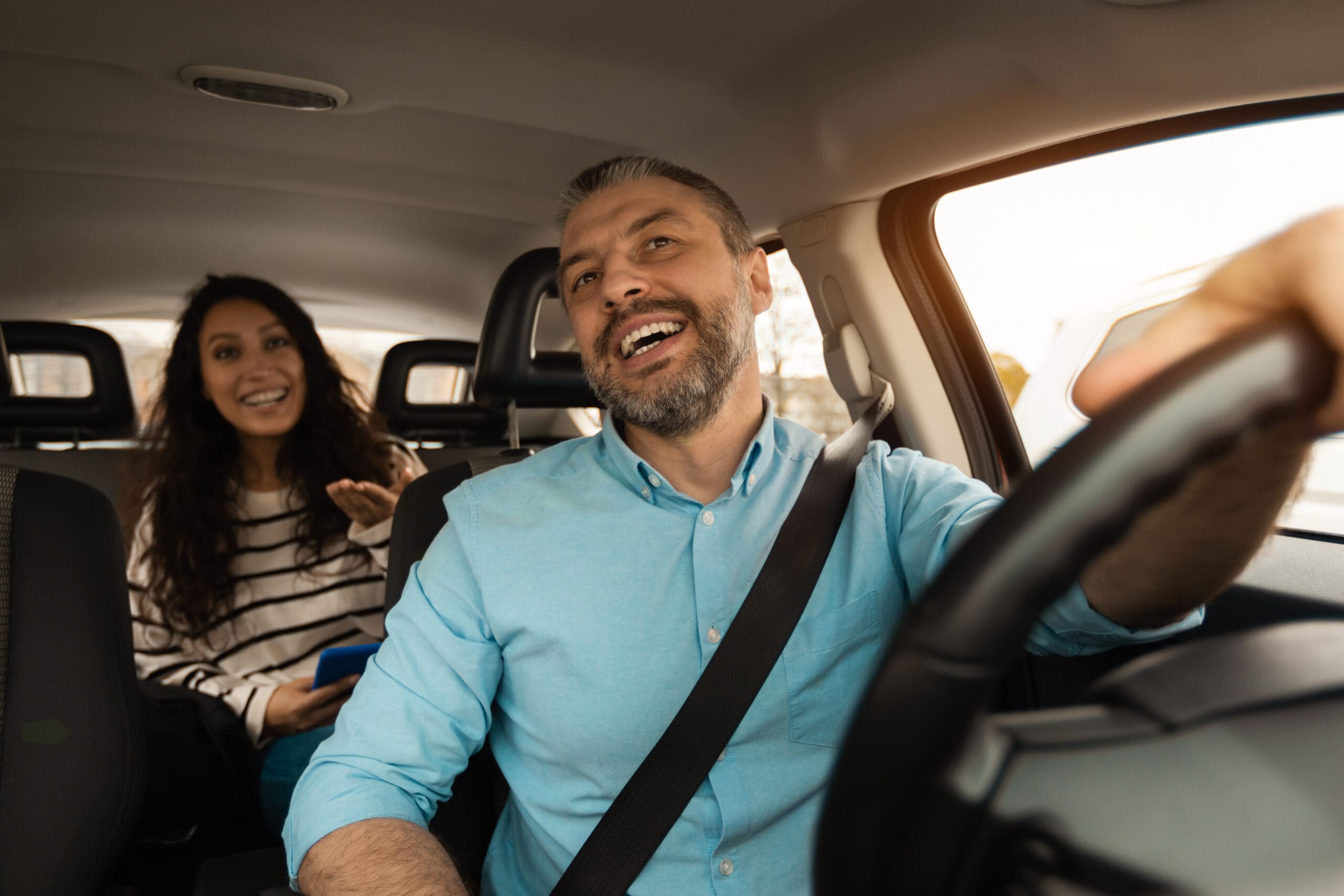 Nice Ride. Portrait of happy male driver riding car looking at cheerful beautiful lady sitting inside auto on back passenger seat, female using cell phone and talking with guy, windshield view