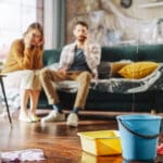 Roof is Leaking or Pipe Rupture at Home: Panicing Couple In Despair Sitting on a Sofa Watching How Water Drips into Buckets in their Living Room. Catastrophe, Distaster and Financial Ruin