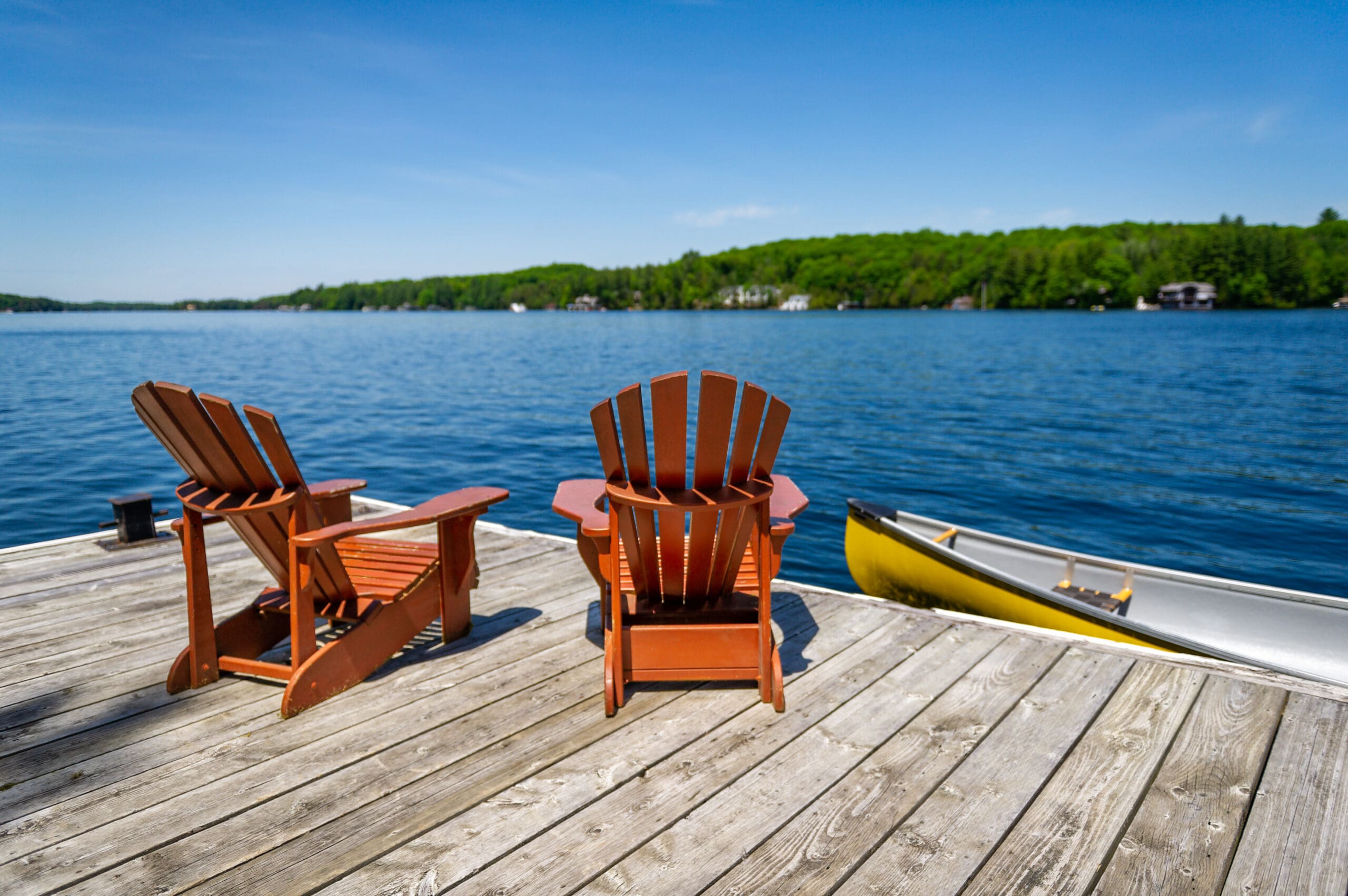 Two Adirondack chairs on a wooden dock facing the blue water of a lake in Muskoka, Ontario Canada. A yellow canoe is tied to the jetty.