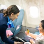 Onboard product purchasing, Asian woman sitting in airplane buying gift from air hostess and paying by credit card tapping on machine. Contactless payment and technology inflight service concept.