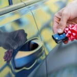 Closeup male hand of new black car is holding gift, surprise, keys with red bow. Lucky driver is going to sitting behind steering wheel of automobile. Man winner won auto in raffle, lottery.