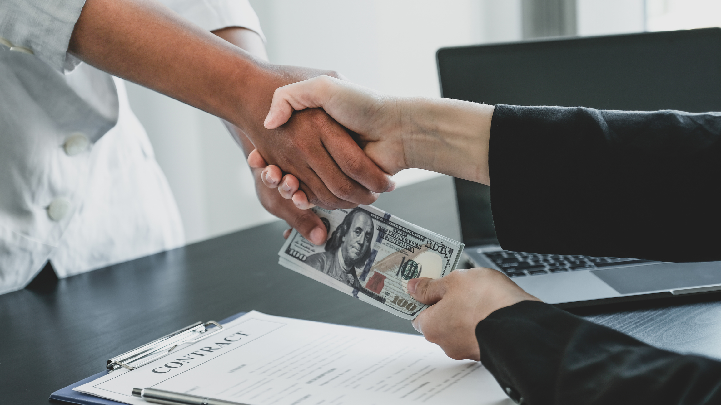 Two hands shaking and exchanging a wad of cash after signing a loan agreement.