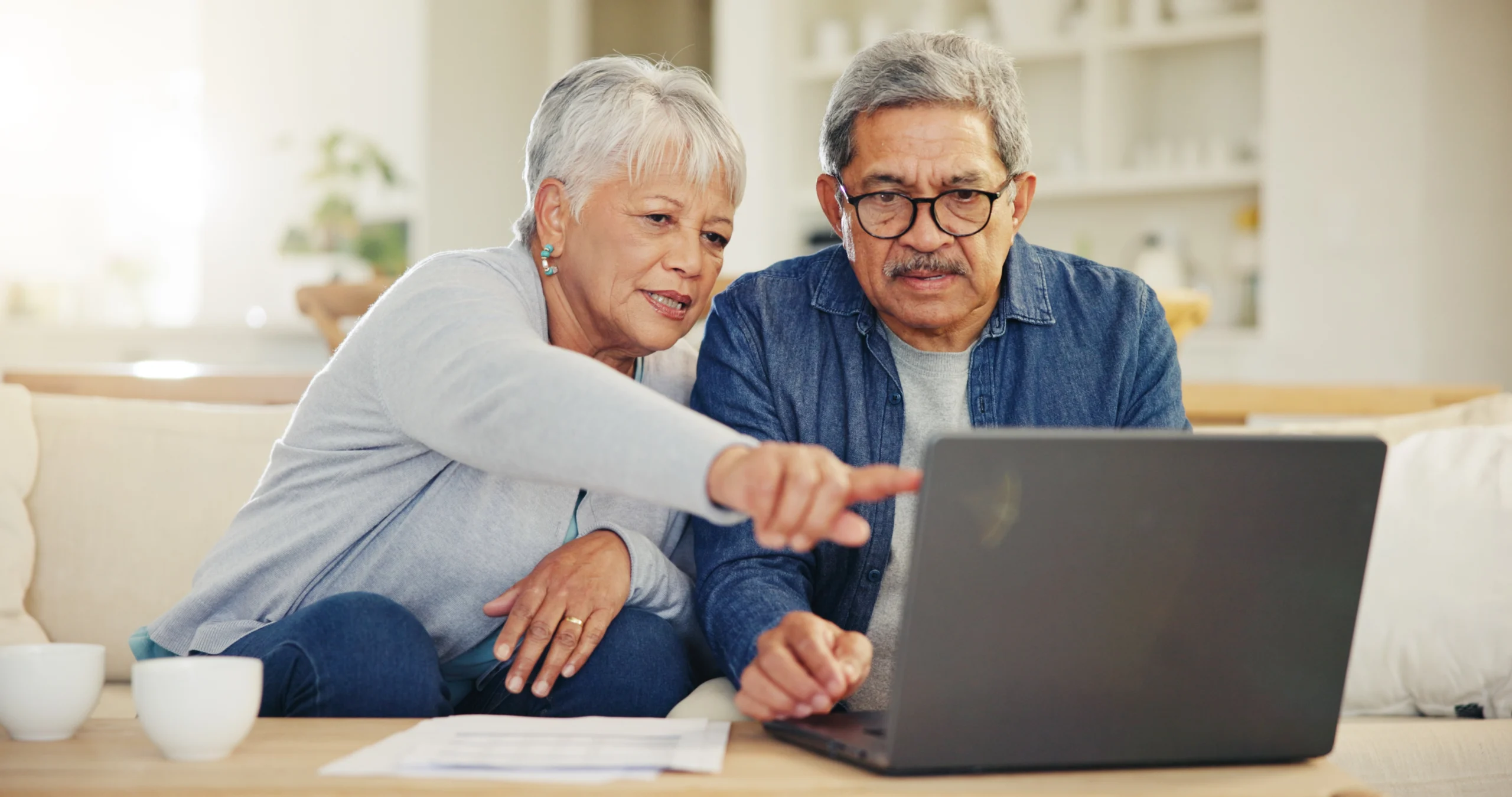 Older Couple Sitting On Couch Looking At Low Risk Investments On Laptop