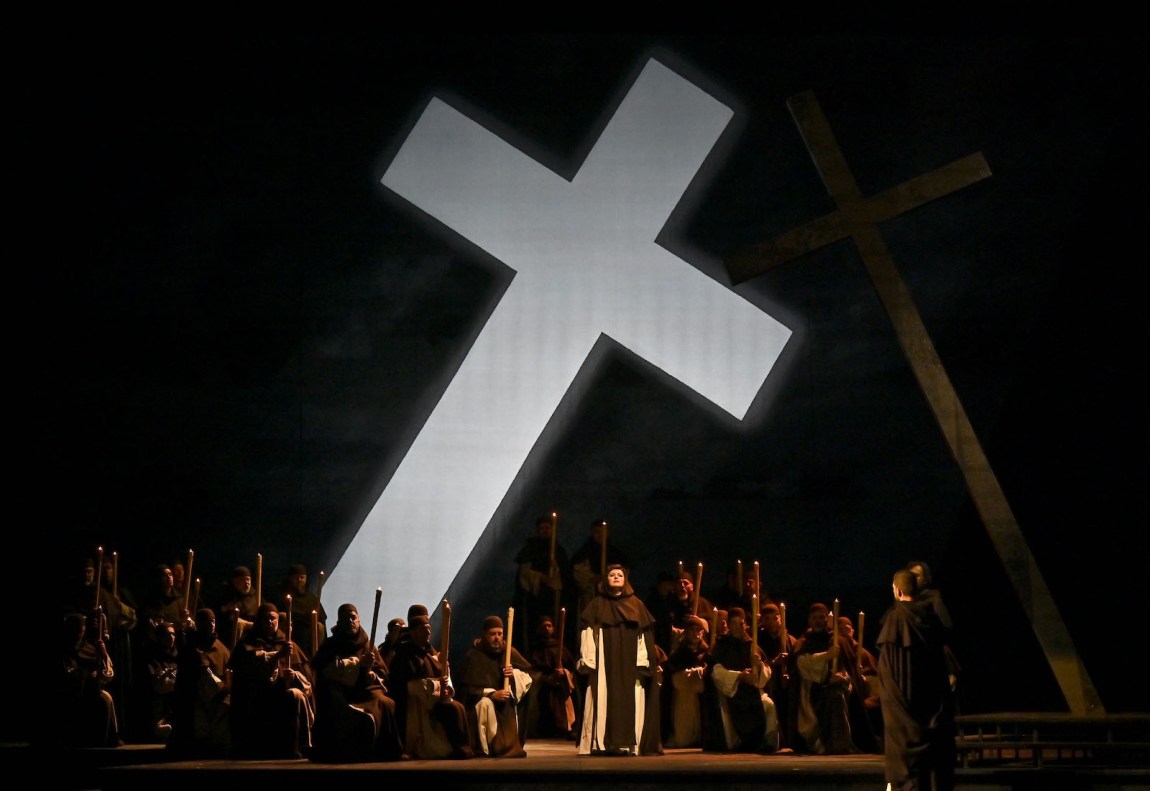 A crowd of monks sings under an enormous, diagonal cross