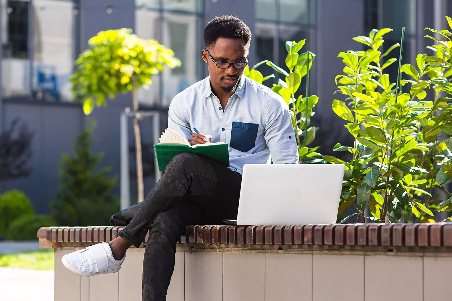 A male student is sitting outside with a notebook and a laptop.