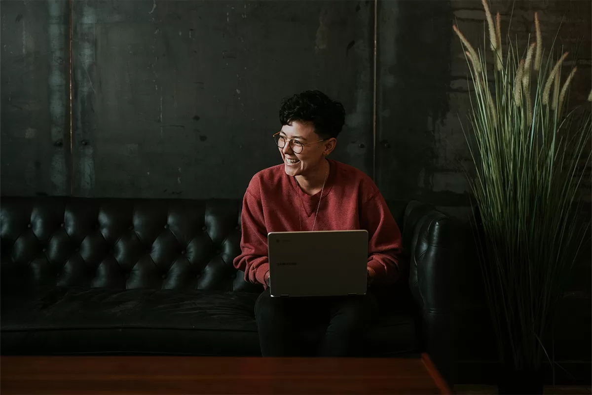 A person sat on a leather sofa with a laptop laughing at something off-camera.