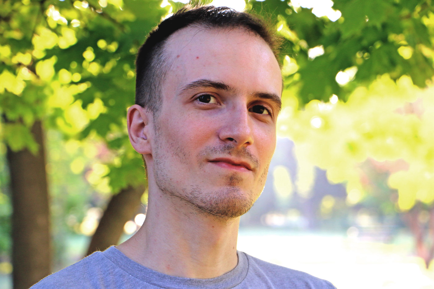 A photo of a light-skinned man with dark hair and a light purple T-shirt looking into the camera with sunlit leaves in the background.