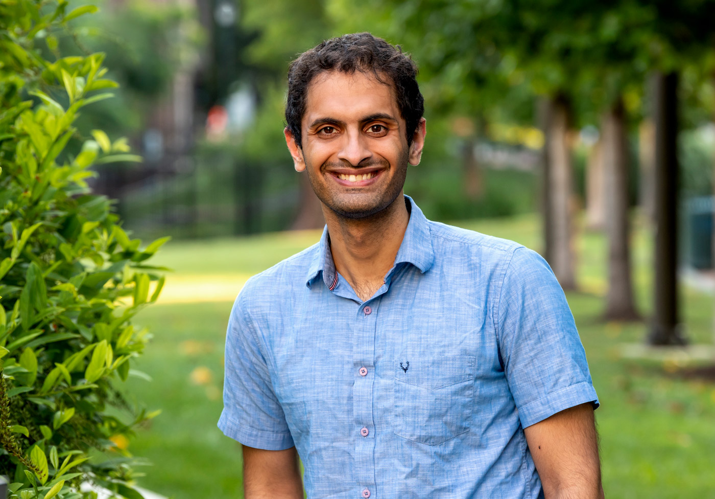 A man with light brown skin smiles into the camera. He's wearing a blue short-sleeved collared shirt and there are trees and grass in the background.