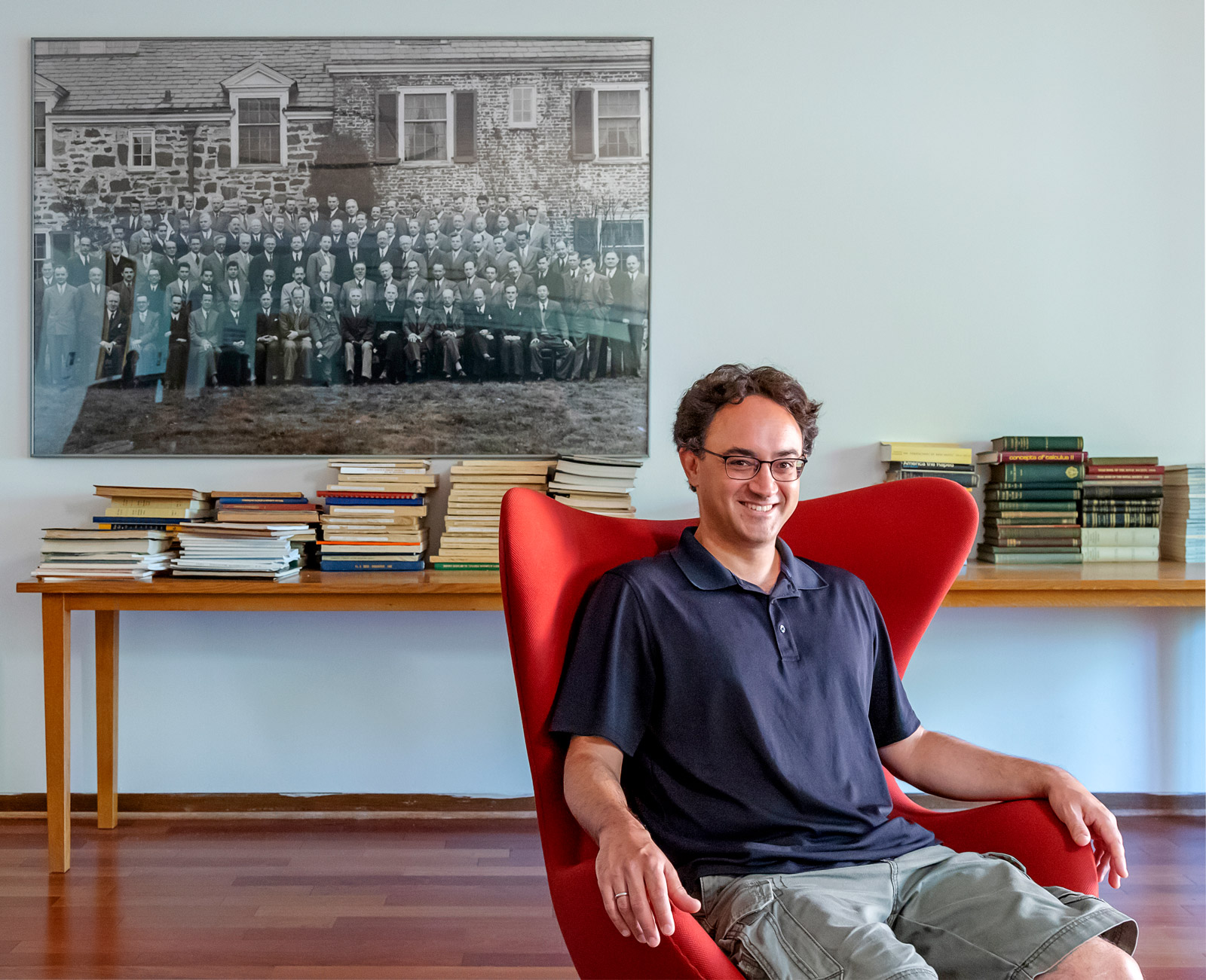 A photo of a dark-haired man in a blue polo shirt sitting in a red wingback chair with books and a large photograph in the background