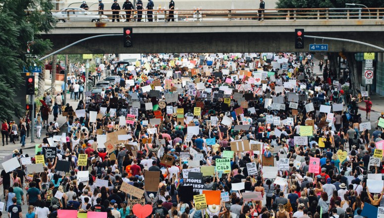 A large group of protesters holding up signs on the streets of Los Angeles, with police officers watching from a bridge above the crowd.