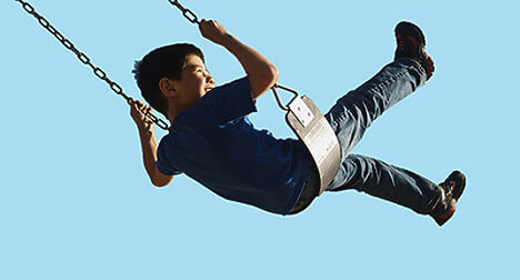 A medium light skinned boy on a swing, mid-swing with feet in the air, against a bright blue background.