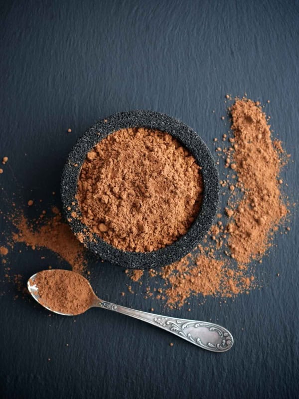 A black bowl filled with cocoa powder sits on a dark surface. Some powder has spilled onto the surface beside it. An ornate silver spoon lies nearby with a bit of cocoa on it.