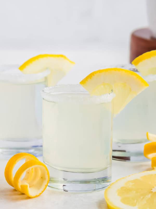 Three glasses of lemon drop shots, garnished with lemon slices on the rims. The glasses have sugared edges and are placed on a white surface, alongside lemon slices and twists. A blurred background with a wooden element is visible.