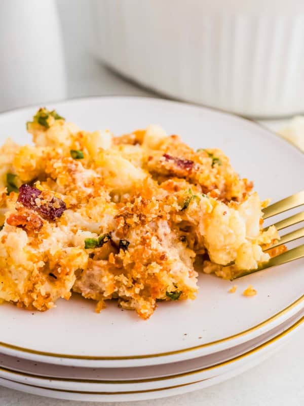 A plate of baked cauliflower gratin with a golden breadcrumb topping, garnished with herbs and small pieces of bacon, placed on a stack of white plates. A fork rests to the side on the plate.