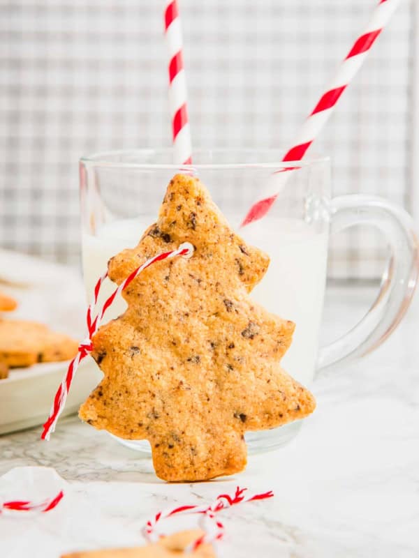 A Christmas tree cookie standing up against a glass of milk with two straws in it.