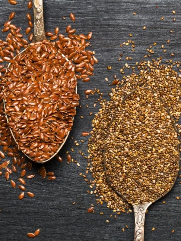 Two spoons filled with flaxseeds and chia seeds rest on a dark textured surface. The flaxseeds occupy the left spoon, displaying a reddish-brown hue, while the chia seeds on the right are smaller and speckled with beige and brown tones.