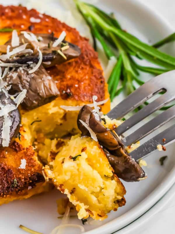 A close-up of a fork holding a piece of golden brown polenta cake topped with grated cheese and mushrooms. The plate is garnished with rosemary, adding a touch of green to the dish.