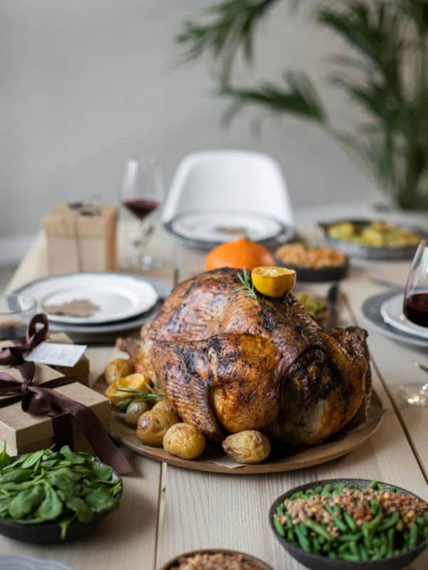 A beautifully roasted turkey on a platter is surrounded by potatoes, offering a nod to Thanksgiving feast alternatives. The table is set with plates of fresh greens, green beans, and grain salad. Glasses of red wine and a wrapped gift box add to the festive atmosphere.