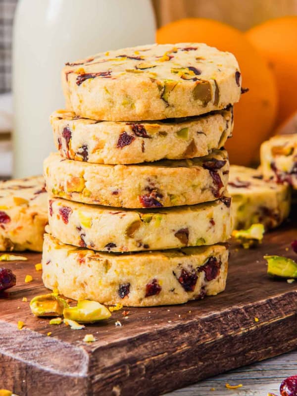 A stack of homemade shortbread cookies with chunks of cranberries and pistachios on a wooden board. In the background, blurred oranges and a bottle of milk are visible. Crumbs and scattered pistachio pieces surround the cookies.