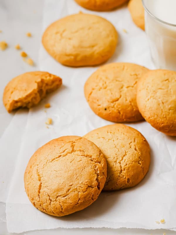 Freshly baked cookies on white parchment paper, one with a bite taken out. A glass of milk is placed nearby. Crumbs are scattered around, giving a homely and inviting appearance.