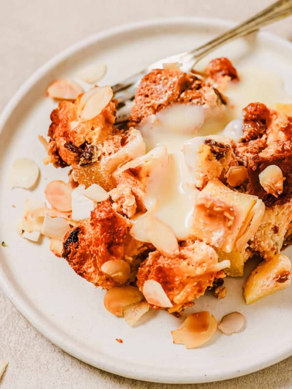 A close-up of a bread pudding dessert on a white plate, featuring a baked mixture topped with custard and sliced almonds. A fork rests on the plate, surrounded by a variety of golden-brown and pale layers.