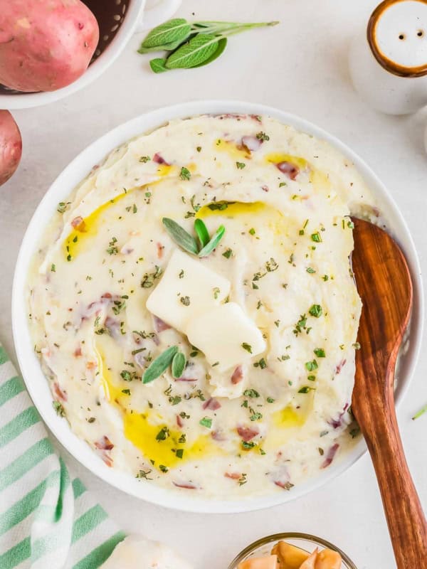 A bowl of creamy mashed potatoes with roasted garlic topped with melted butter, herbs, and a sprig of sage. A wooden spoon rests in the potatoes. Fresh red potatoes, a striped cloth, and seasoning dishes are nearby.