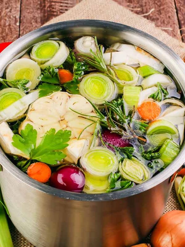 A pot filled with fresh vegetables including leeks, carrots, garlic, celery, and herbs, sits on a wooden surface. Additional vegetables and herbs are scattered around the pot.