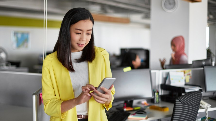 Female office worker, professionally dressed in a bright yellow jacket, stands near her desk while she checks her employee self-service system from her smartphone