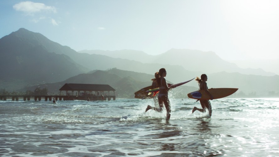 Three people on a beach, running into the ocean with surboards in a tropical climate, with mountains in the background.