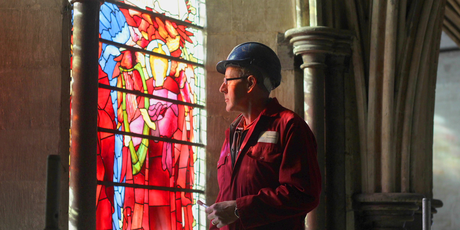 Man in overalls and a hat hat inspecting a stained glass window