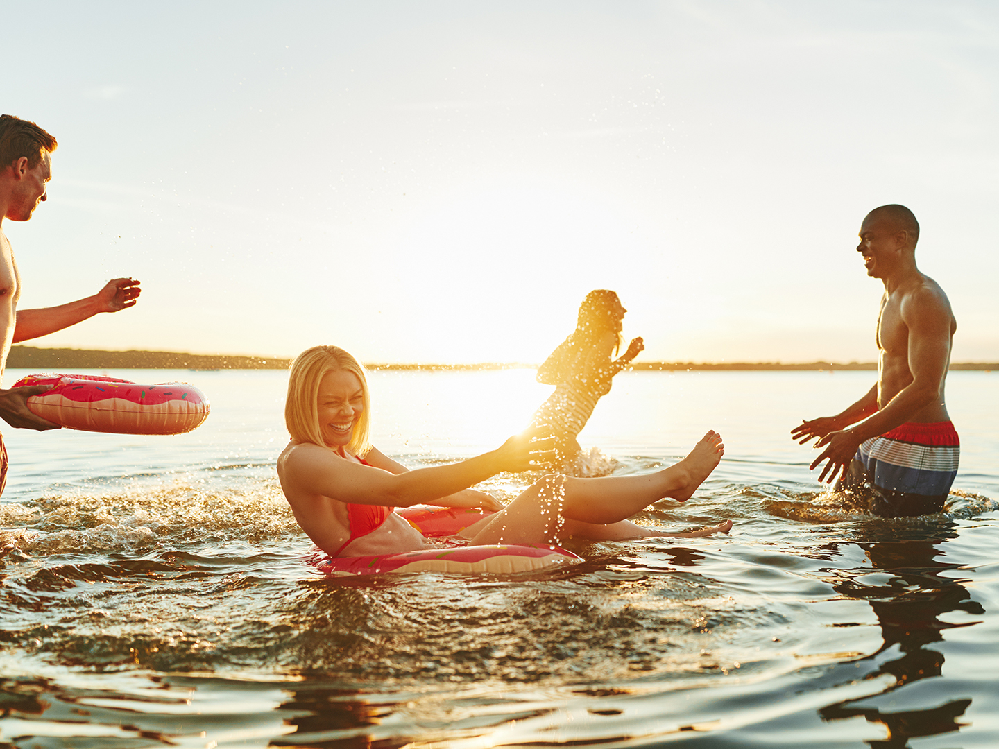 Diverse group of young friends in swimsuits laughing and splashing each other while having fun together in a lake at sunset