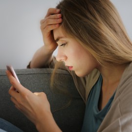 Portrait of young woman with depressed facial expression sitting on grey textile couch holding her phone.