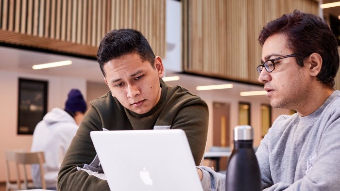 Two students working from the same laptop at a table in the City Campus atrium