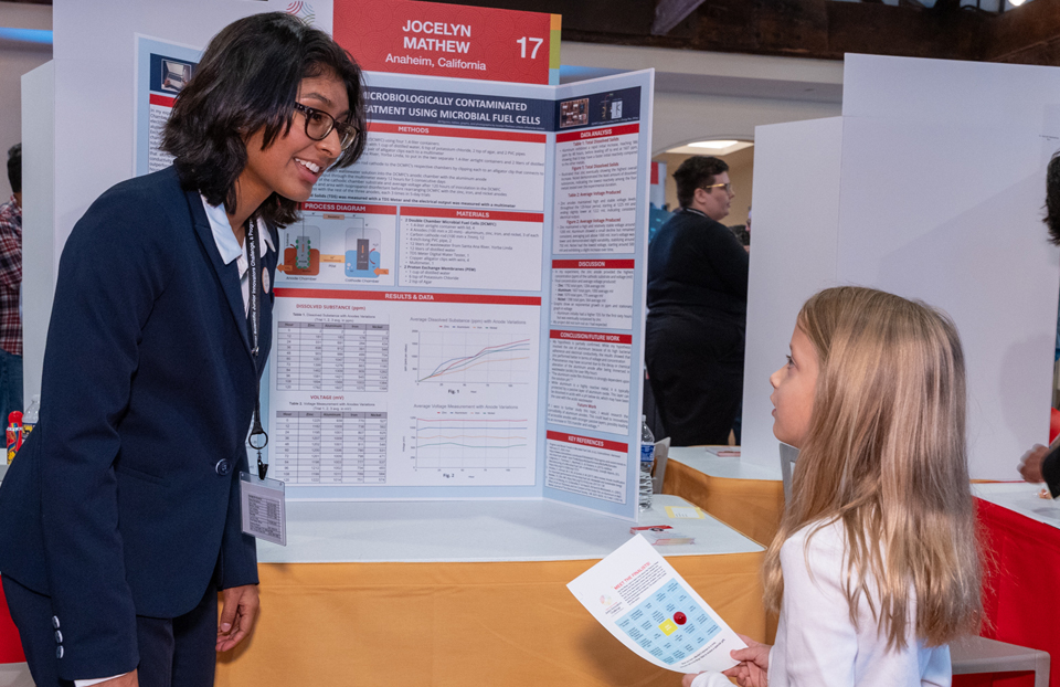 Thermo Fisher Top 30 finalist talks to Public Day guest. The finalist, has short black hair and wears glasses and is wearing a blue suit. The Public Day guest, is wearing white and has blonde hair.