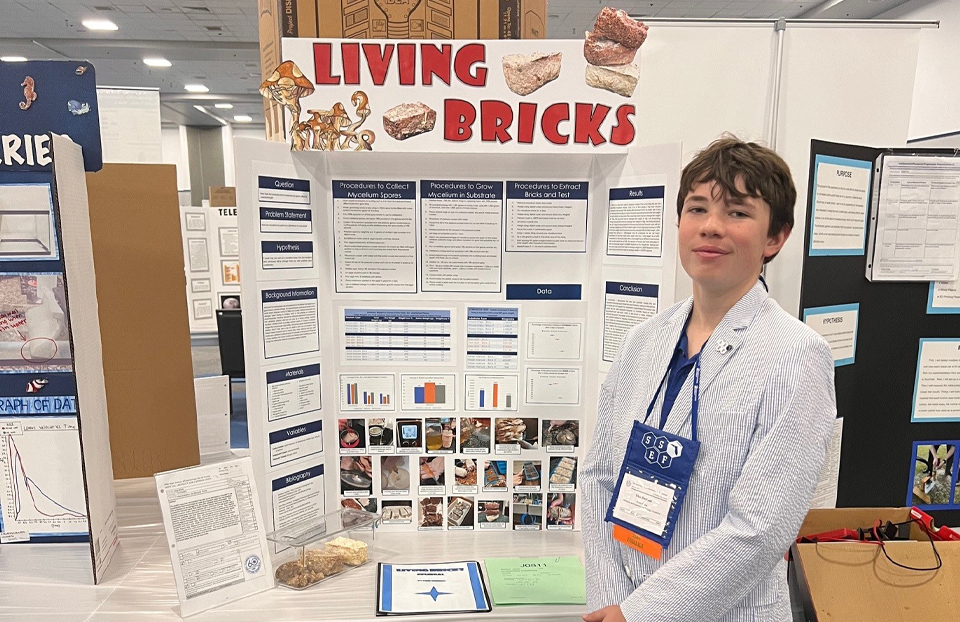 A boy with brown hair poses in front of his project board.