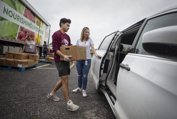 Two people load a cardboard box into the sliding door of a parked white van. More boxes are behind them.