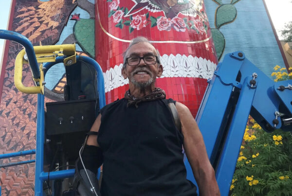 A man stands in front of An older man stands in front of a mural he just painted. He's grinning and facing the camera, wearing glasses.